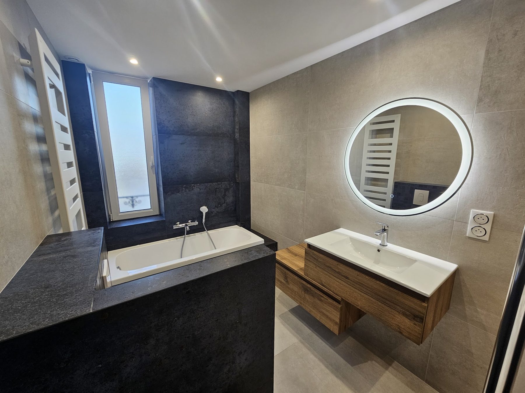 Salle de bains en pierre anthracite, baignoire, miroir rond lumineux et meuble en bois